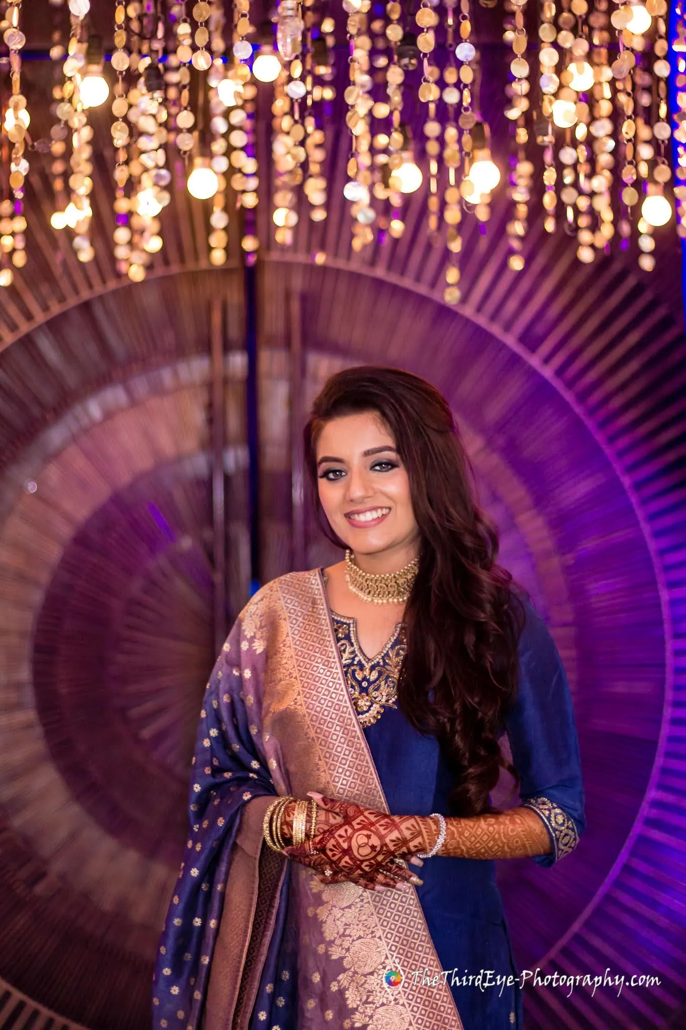 A woman in a blue embroidered outfit with henna stands before a round gold backdrop and crystal lights