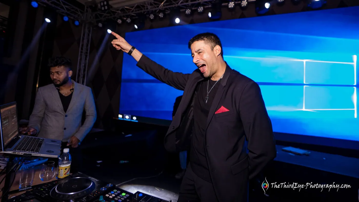 A man in a black jacket points upward and sings out beside the DJ desk under a blue screen