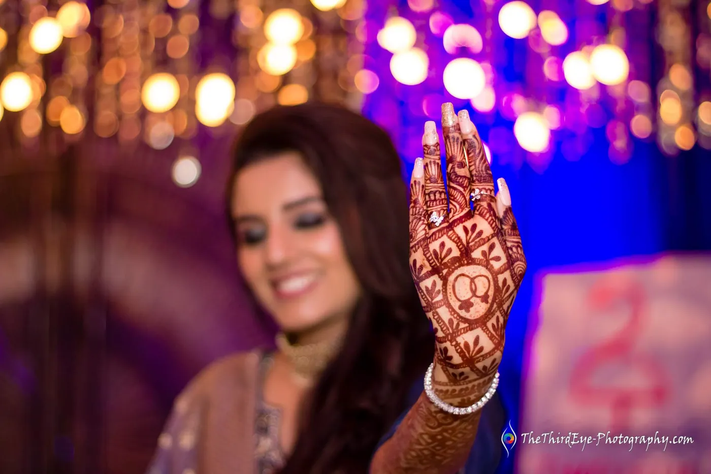 A woman smiles softly behind a henna covered hand raised toward the camera, golden lights glowing behind