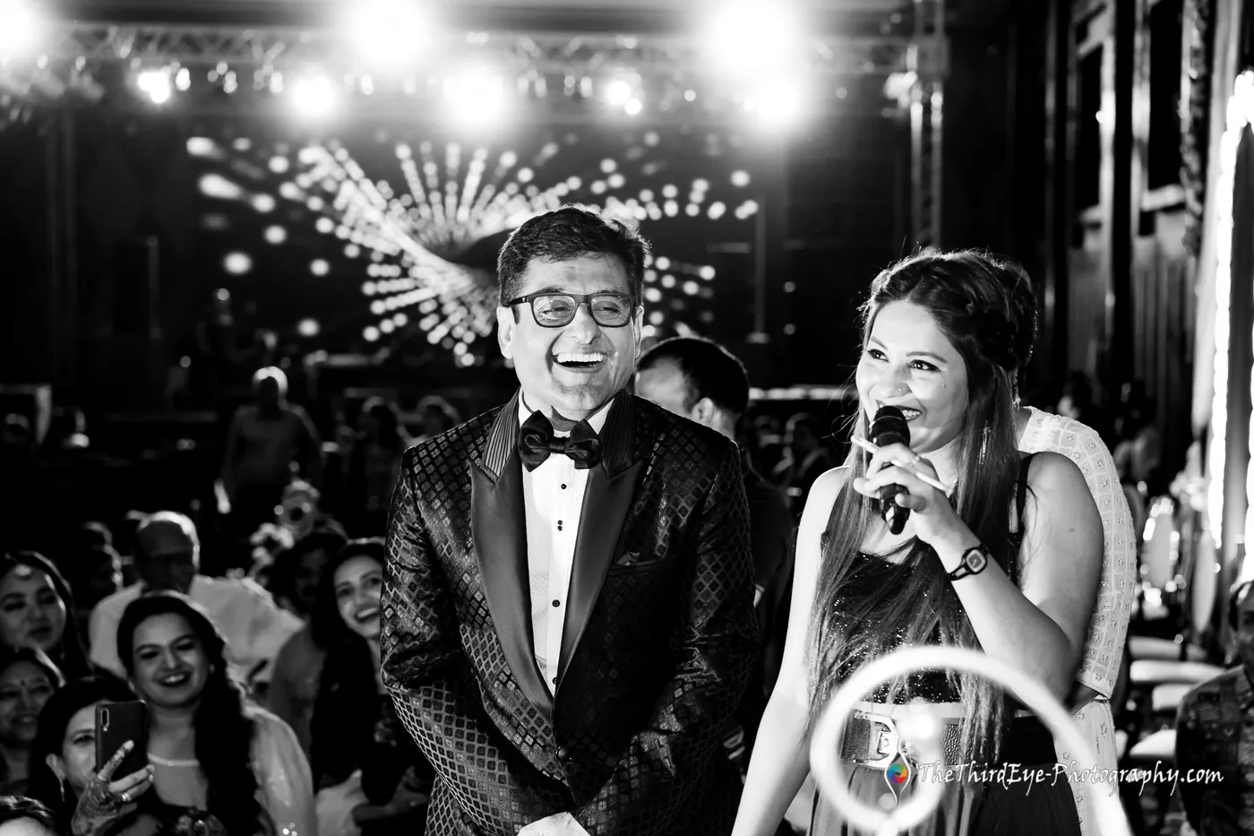 Black and white frame of a man in a tuxedo and a woman with a microphone laughing before a crowd