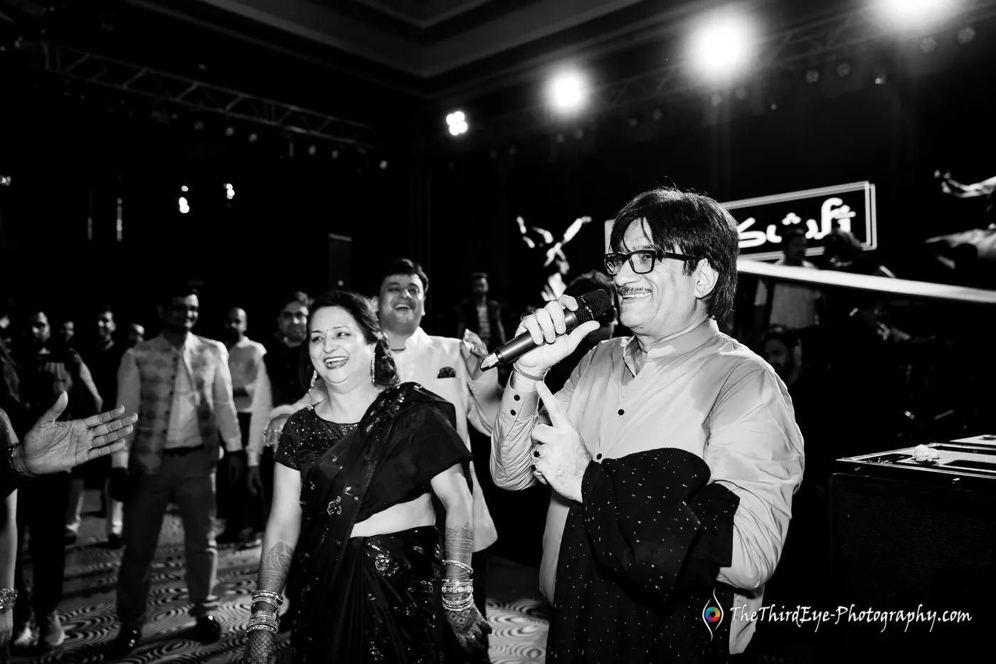 Black and white frame of a woman with a microphone and a man in glasses performing in a dim crowd