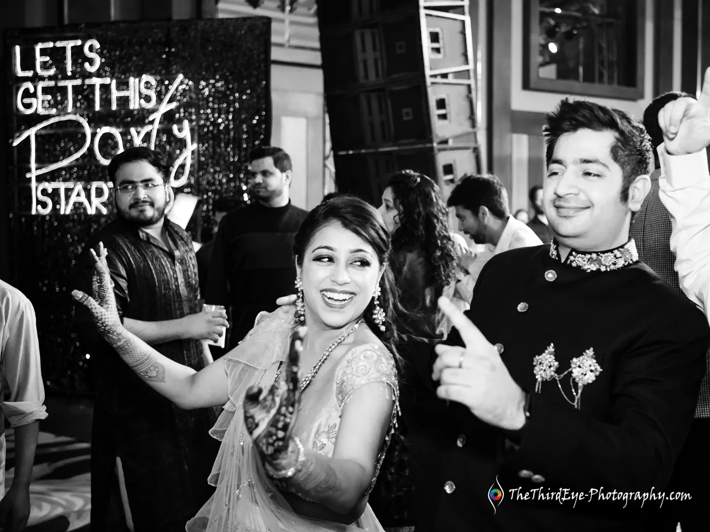A woman with a braid laughs and dances beside a man in a dark sherwani under a glowing party sign