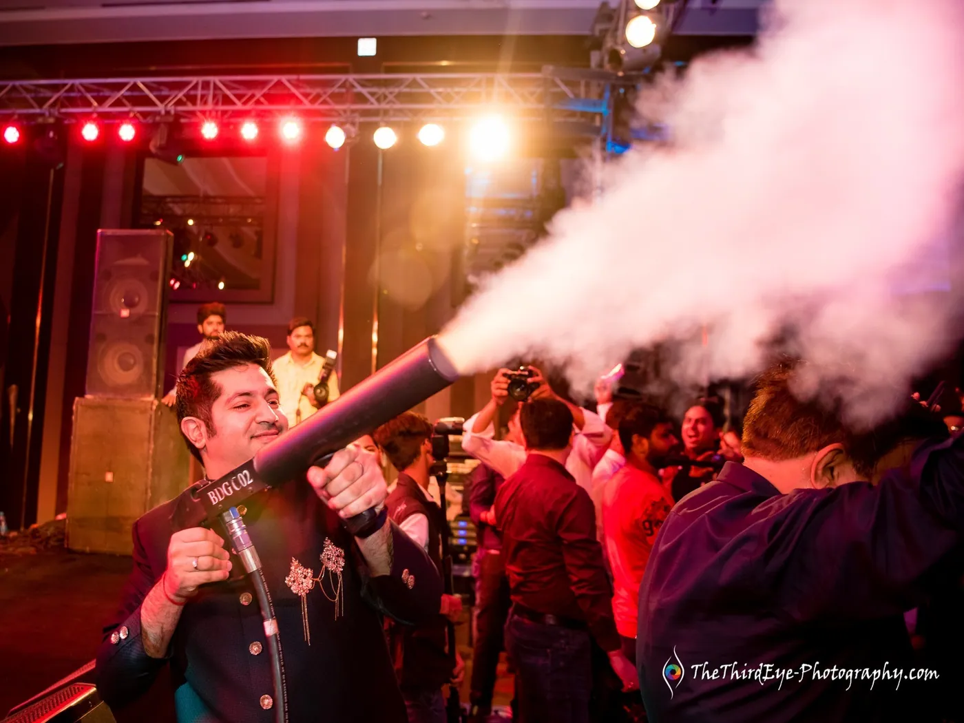 A man in a black jacket fires a cannon of white smoke over the dance floor under red lights