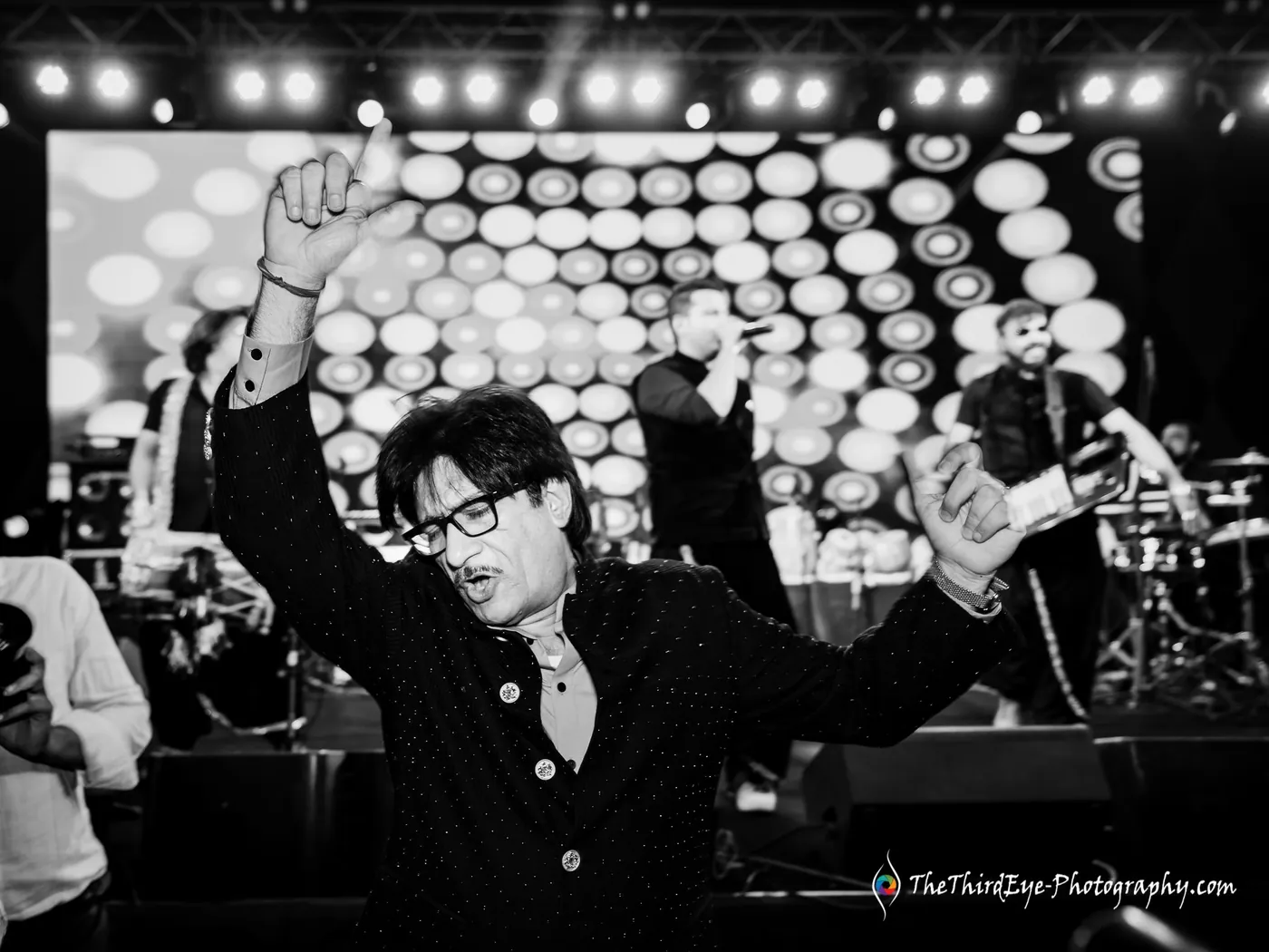 Black and white man in glasses and a dark jacket throws one arm into the air on the dance floor