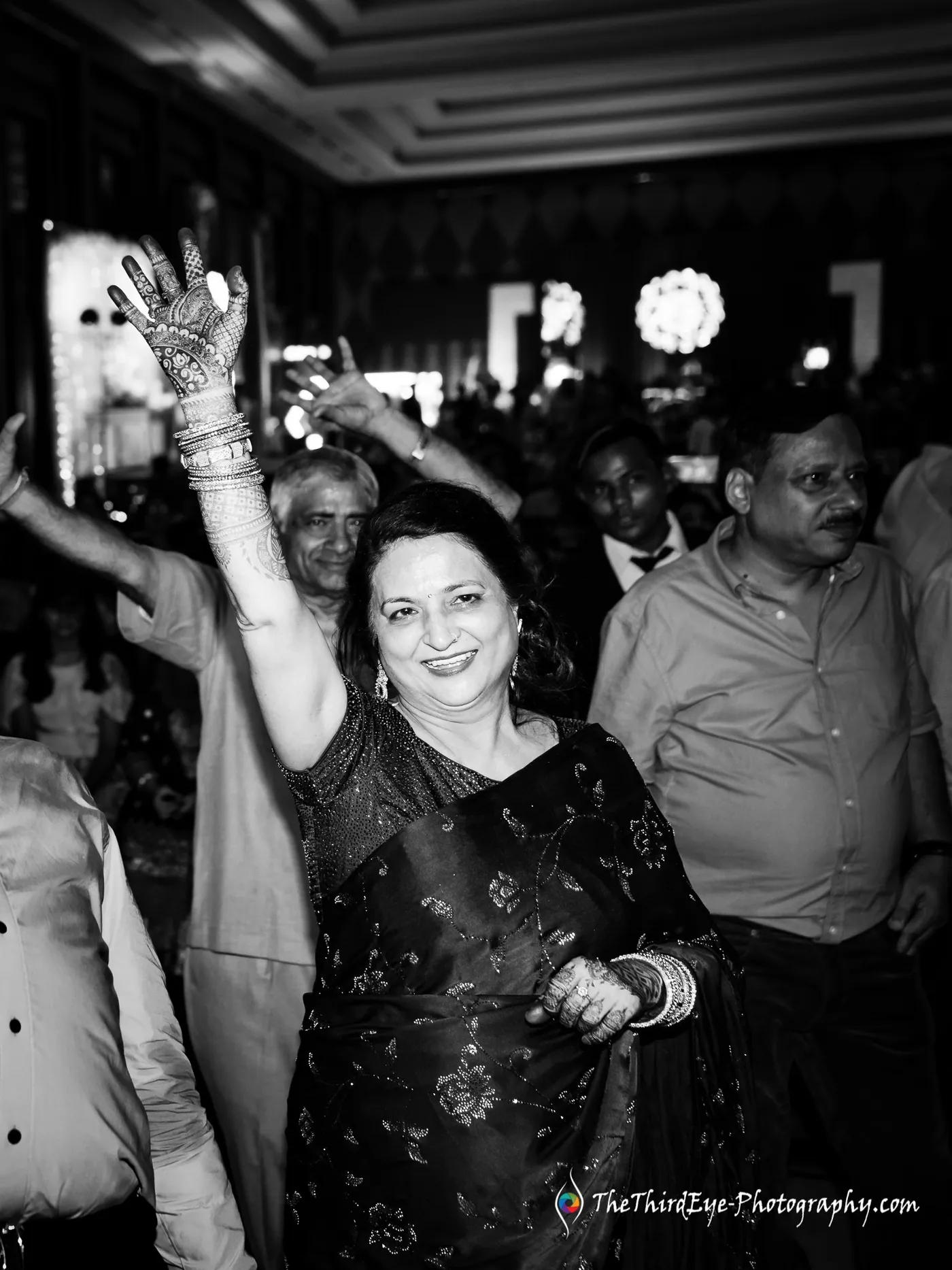 Black and white frame of a woman with a raised henna painted hand dancing in a crowd, smiling