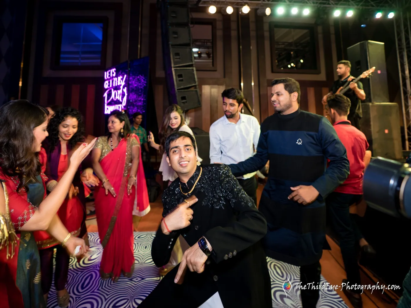 A man in a dark jacket crouches with a pointing gesture while friends dance around him under coloured lights