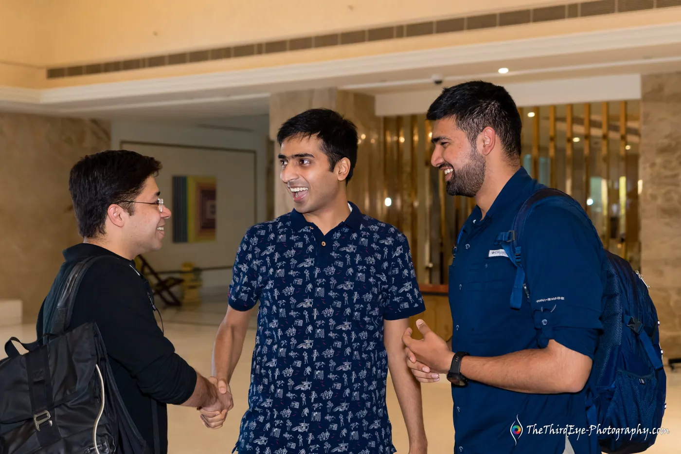 Three men meet in a hotel lobby and shake hands, one carrying a backpack and another in a blue shirt