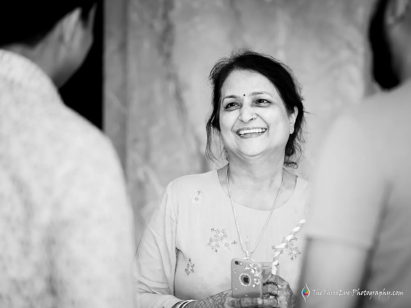 Black and white close frame of an older woman laughing warmly while holding a phone, men's shoulders in the foreground