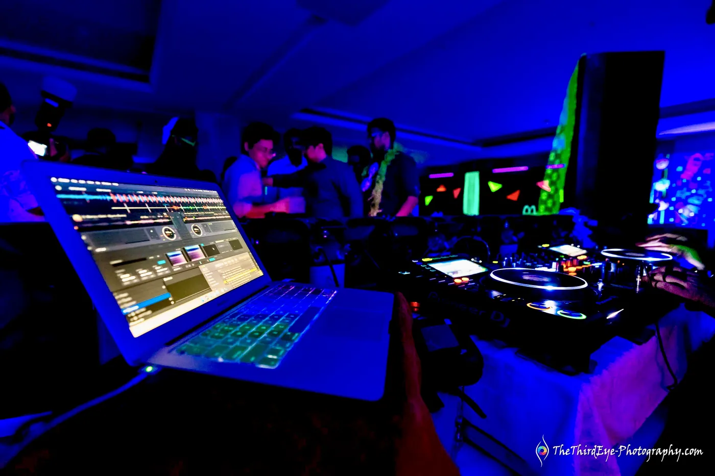 DJ laptop and turntables glow in blue light with guests moving in the dark behind