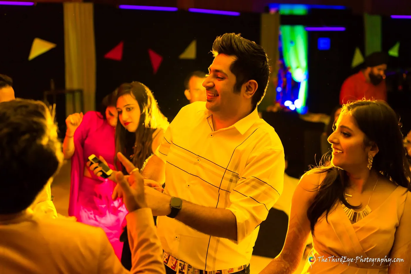 A man in a yellow checked shirt smiles at a phone as friends lean around him in neon light