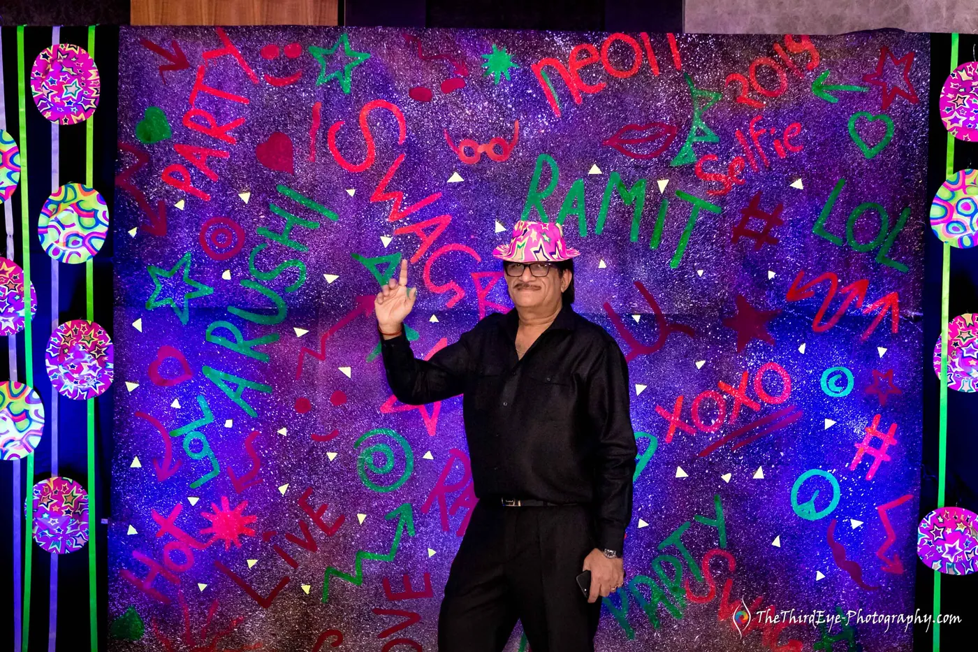 A man in a patterned hat poses with one arm raised before a backdrop of glowing scribbles and names