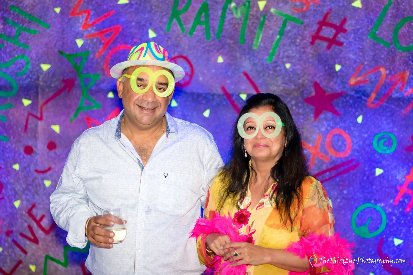 An older man with a drink and a woman in a pink feather boa pose in neon glasses
