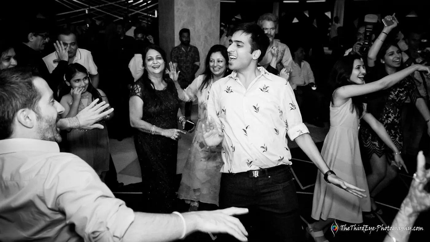 Black and white frame of a man in a patterned shirt dancing with arms out as others dance around him