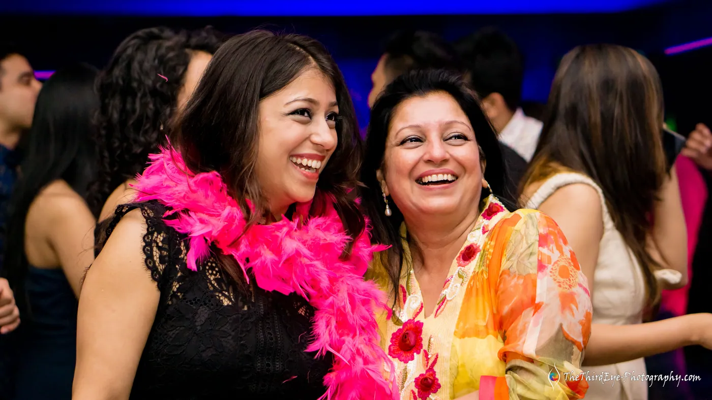 Two women in hot pink feather boas lean together and laugh hard in the dim room
