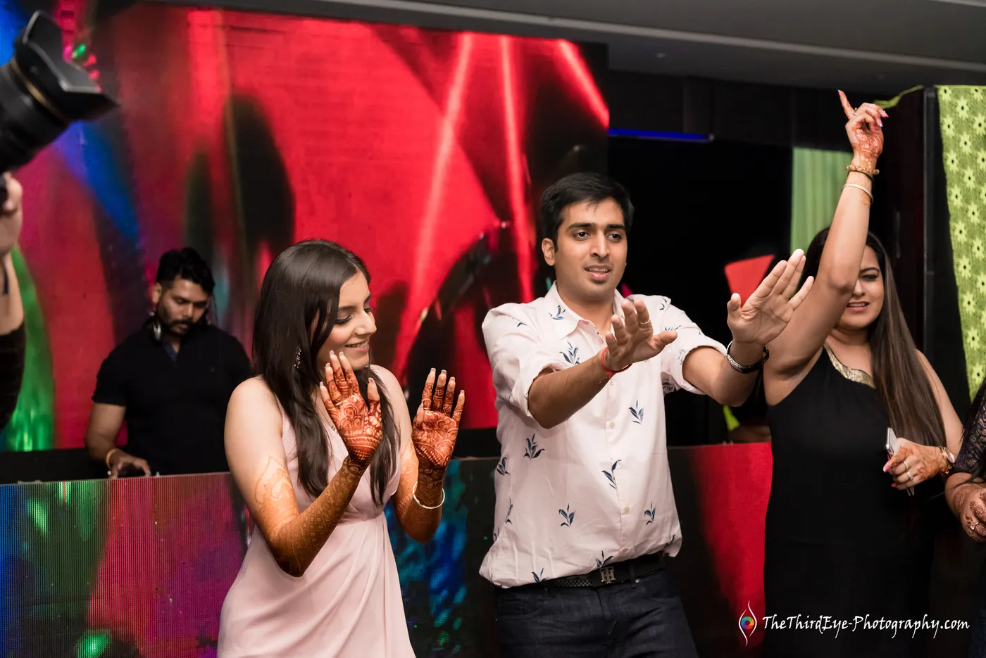 A man and a woman dance with arms flung wide while friends cheer on the lit floor