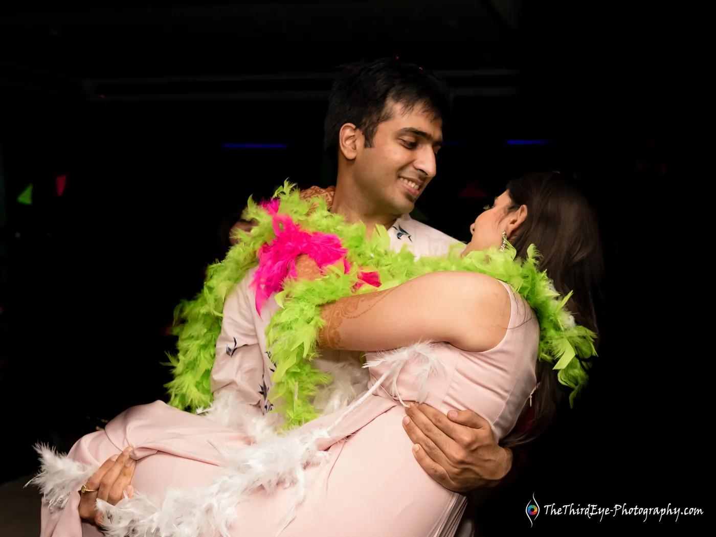 A man sweeps a woman off her feet, both draped in a neon green and pink feather boa