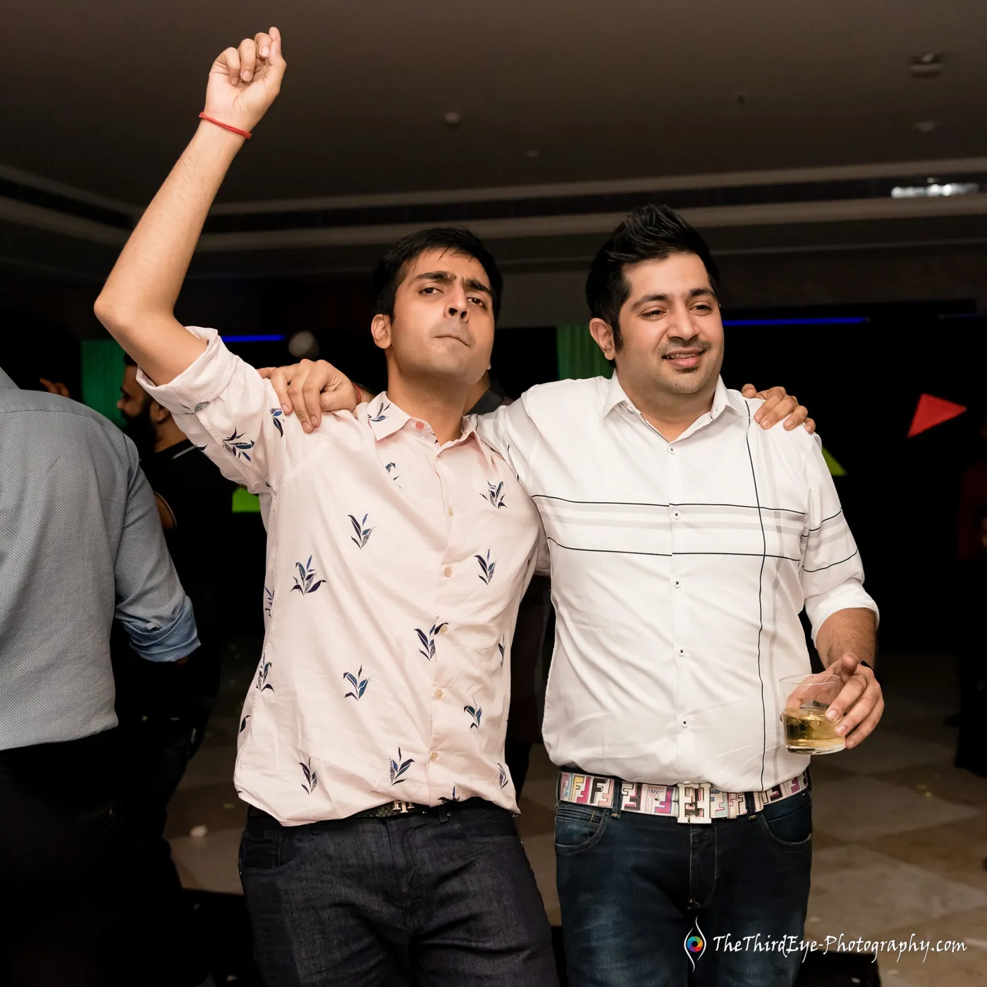Two men in patterned shirts lean in together, one punching an arm high with a drink in his other hand