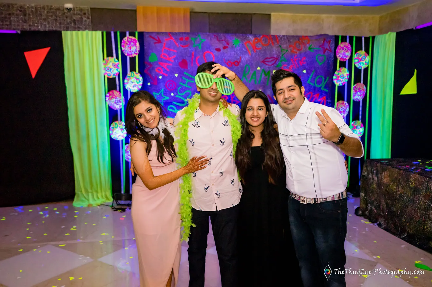 Four friends pose with arms around each other on a dance floor, fluorescent balloons and green panels behind them