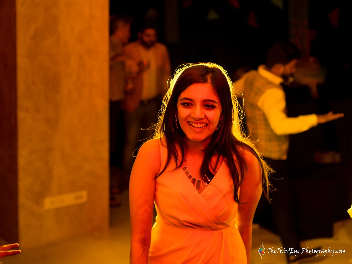 A woman in an orange dress laughs in a warm amber glow with people blurred behind her