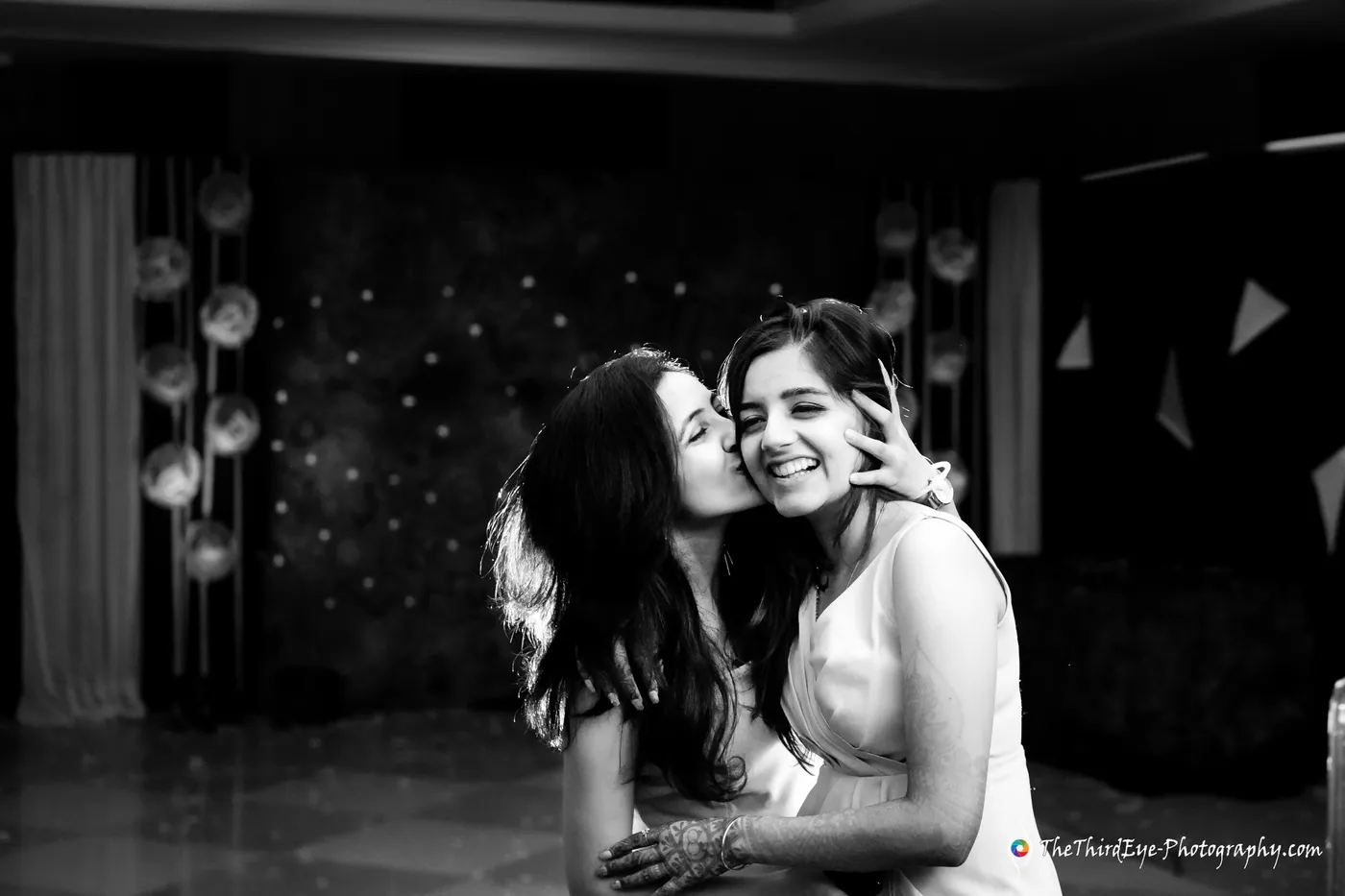 Black and white frame of two women in a tight embrace, one kissing the other's cheek in the dim room