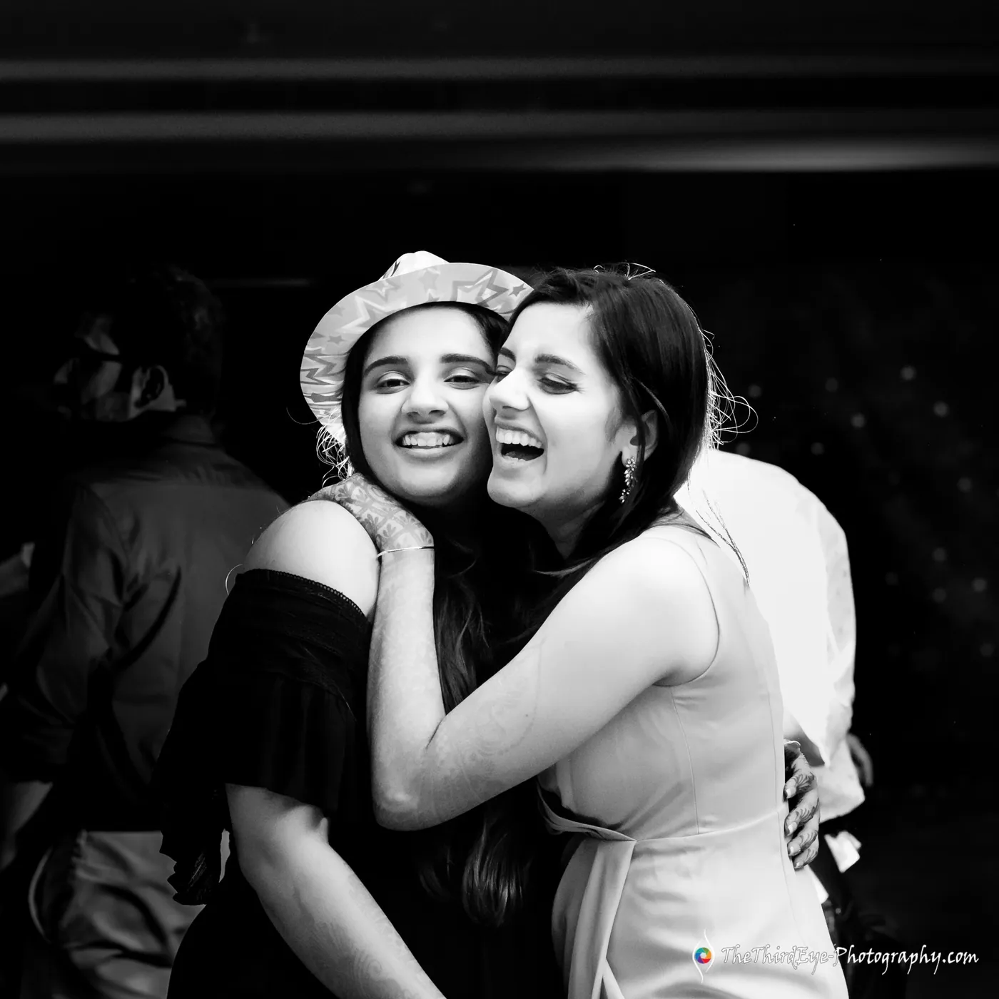 Two women hug and laugh hard with cheeks pressed together, one in a brimmed hat, in black and white