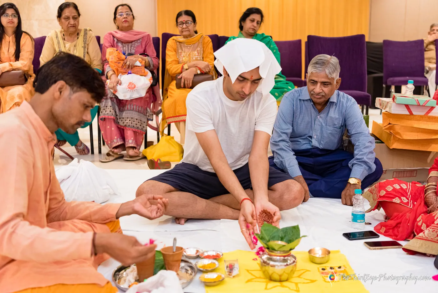 A man in a white head cloth sits cross legged at the haldi beside a priest as elders watch