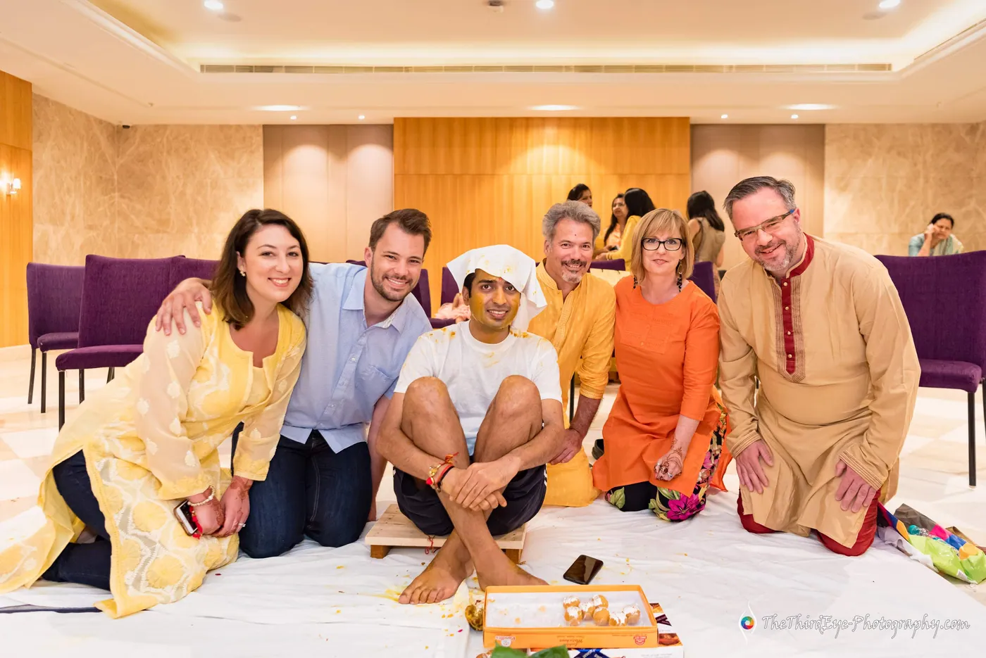 A group sits on white sheets at the haldi, a man in a head cloth among smiling relatives