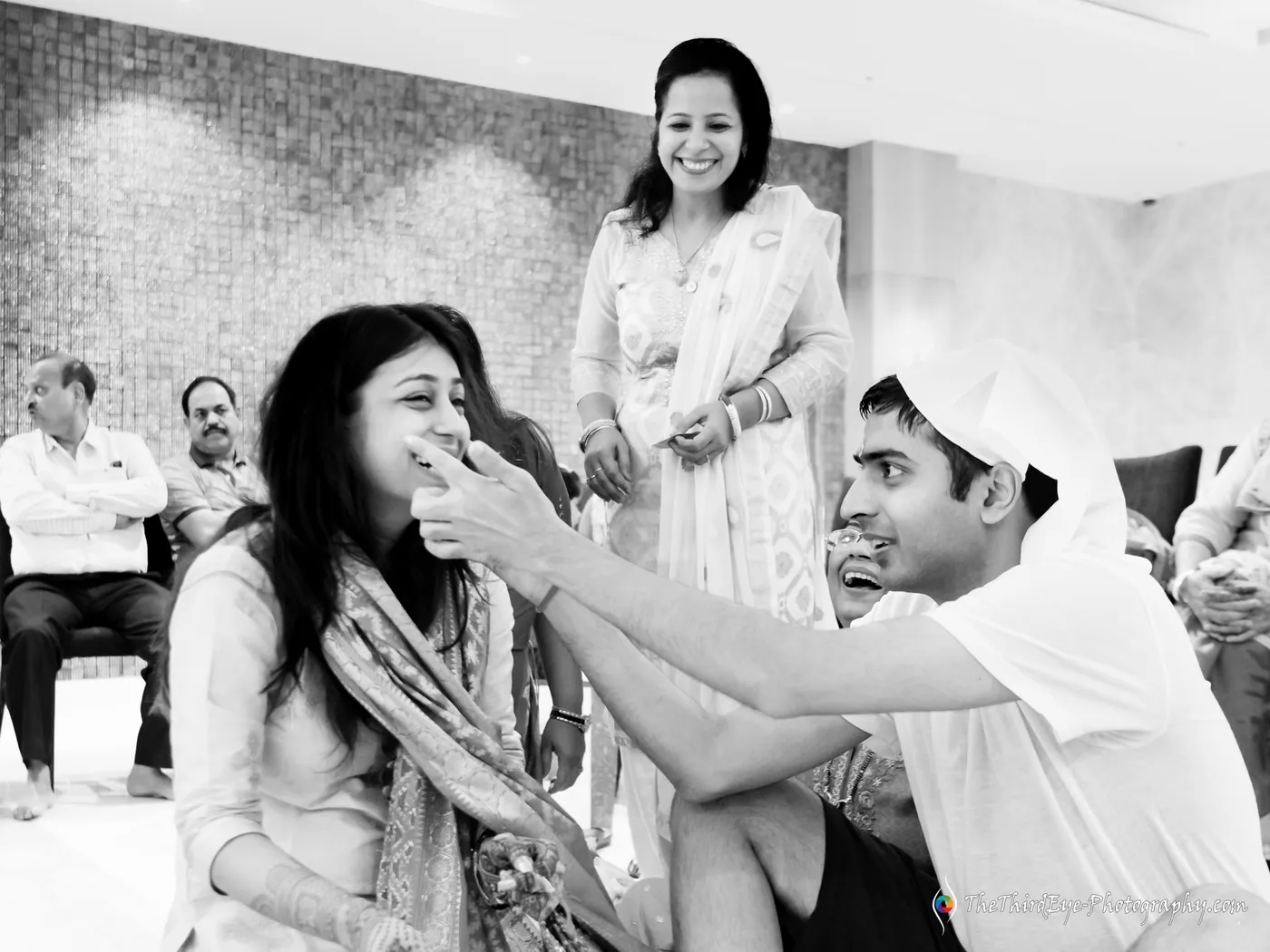 Black and white frame of a man smearing haldi on a woman's cheek as she scrunches her face and laughs