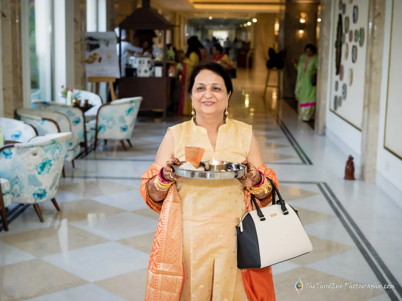 A woman in gold and orange walks down a bright hotel corridor carrying a decorated thali