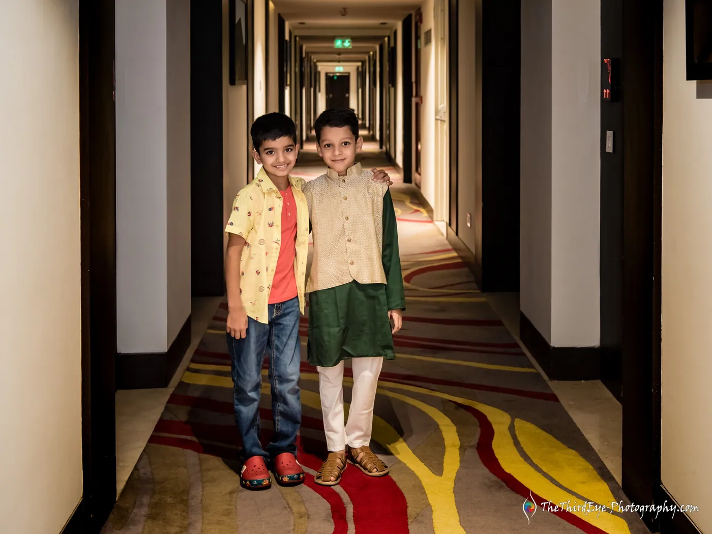 Two boys in kurtas, one yellow and one green, stand side by side in a hotel corridor