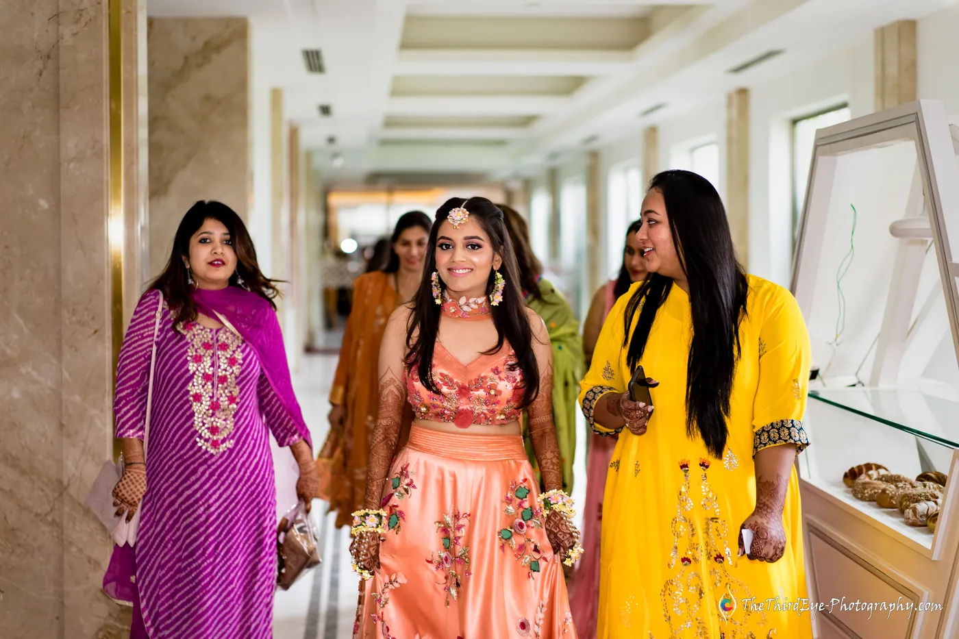 Three women in bright outfits walk toward the camera down a hotel corridor, the middle one in peach