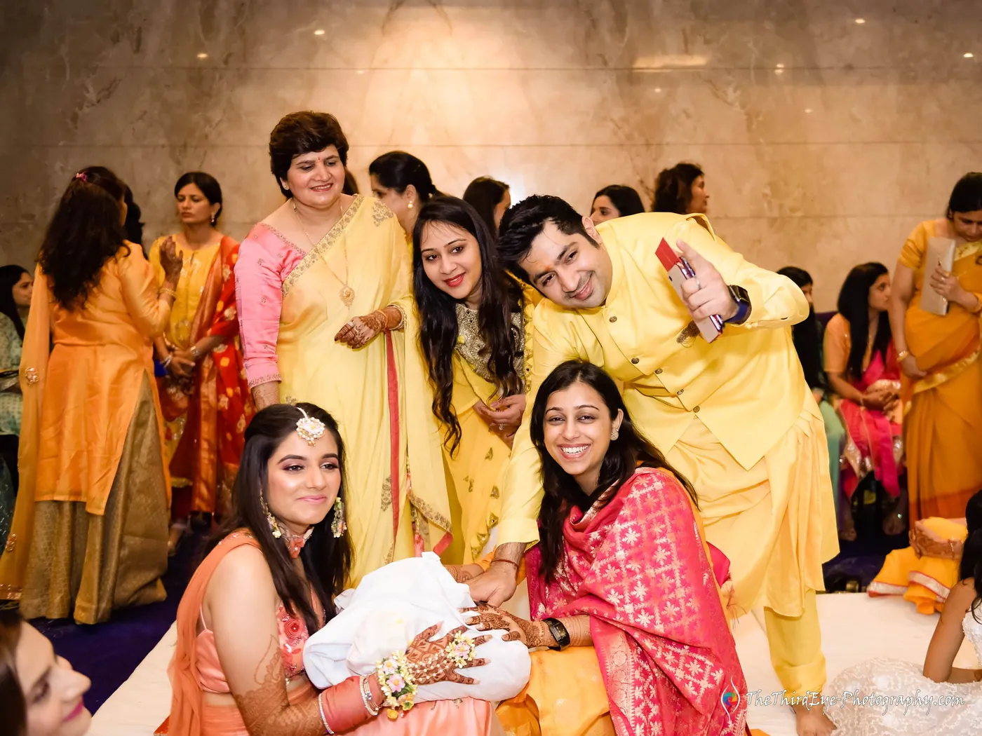 Relatives and a young man in yellow crowd around a woman on the floor, all grinning as haldi is applied