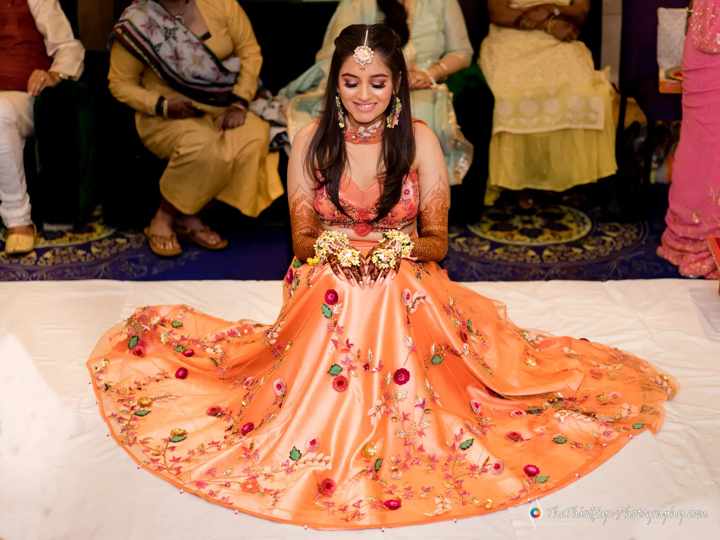 The bride sits on the floor with her embroidered orange lehenga spread around her, smiling down at the haldi seating