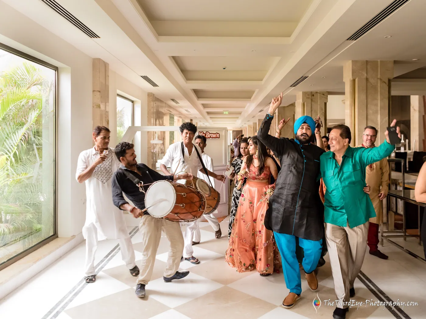 Dhol players lead two dancing men in turbans through the hotel corridor as a crowd follows