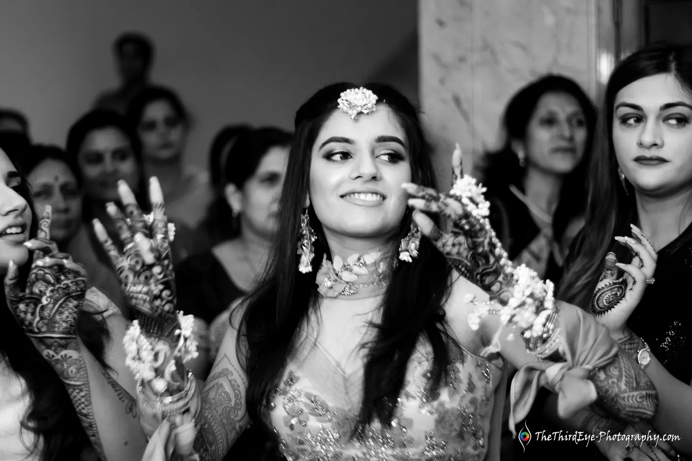 Black and white frame of the bride smiling and showing her henna covered hands as women crowd behind her