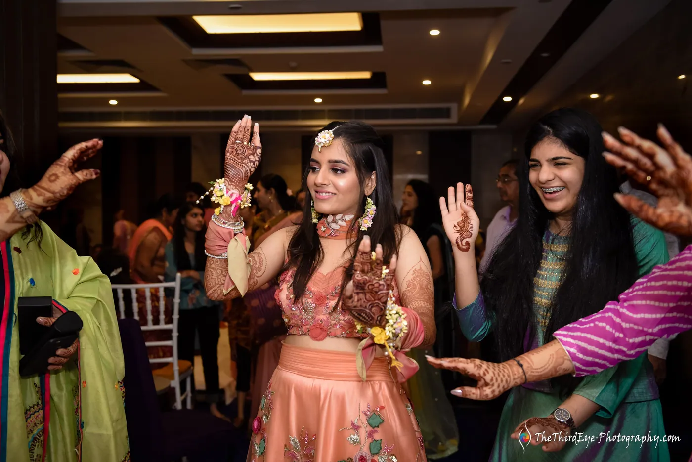 The bride raises a hand wearing a flower bracelet and laughs as women reach in from both sides