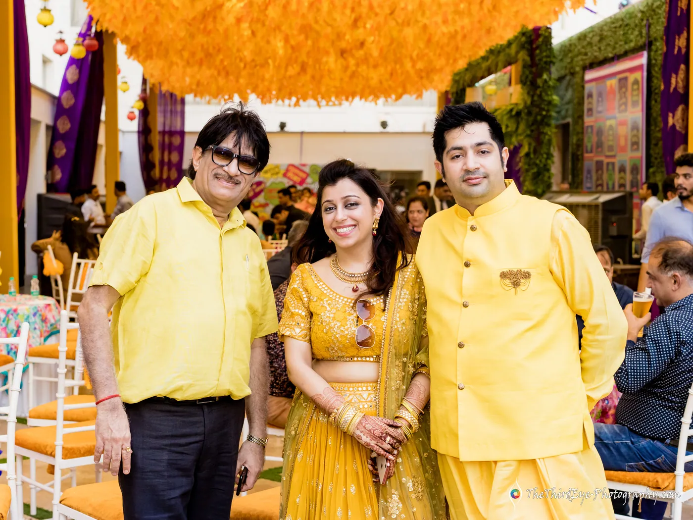 Three people in matching yellow outfits stand together under a ceiling of orange marigolds