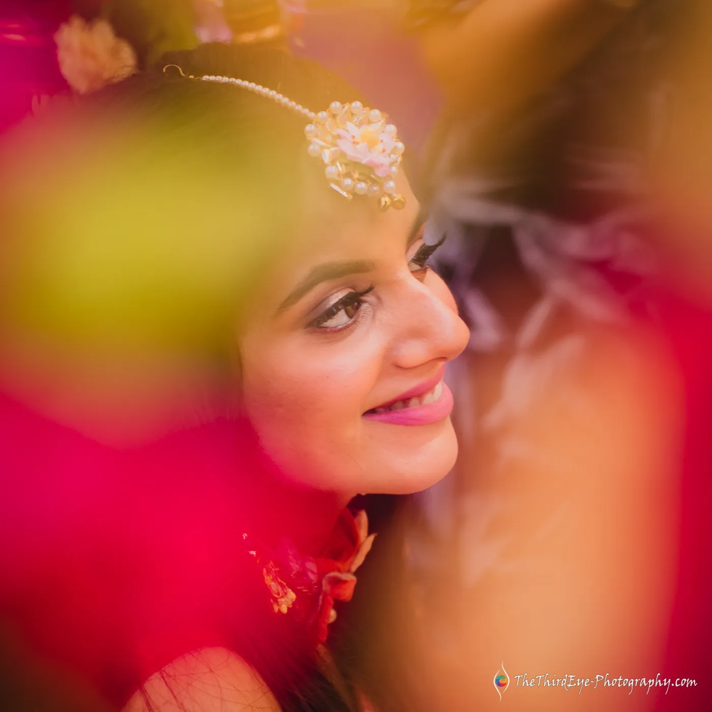 The bride's smiling face seen through a swirl of pink and yellow blur, a gold tikka on her forehead