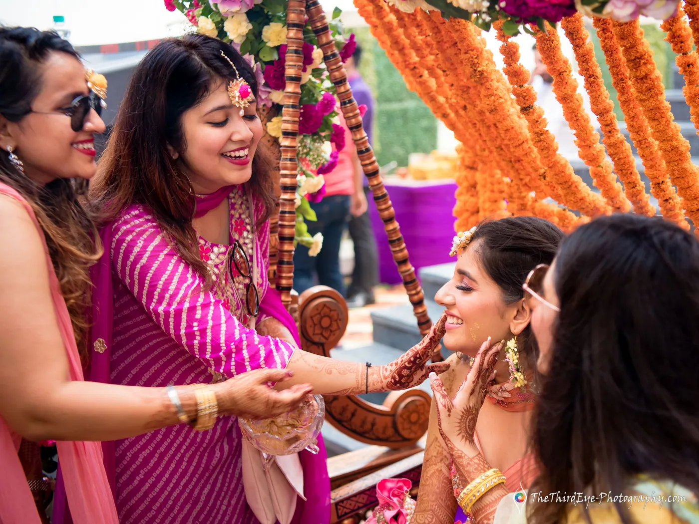 Women gather around the bride on a flower decked swing, one holding her hands as she smiles
