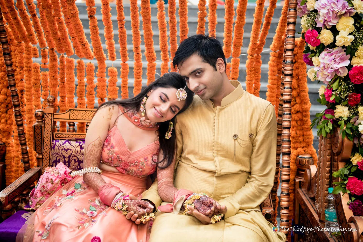 Ramit and Arushi lean their heads together on a flower decked swing in front of marigold garlands