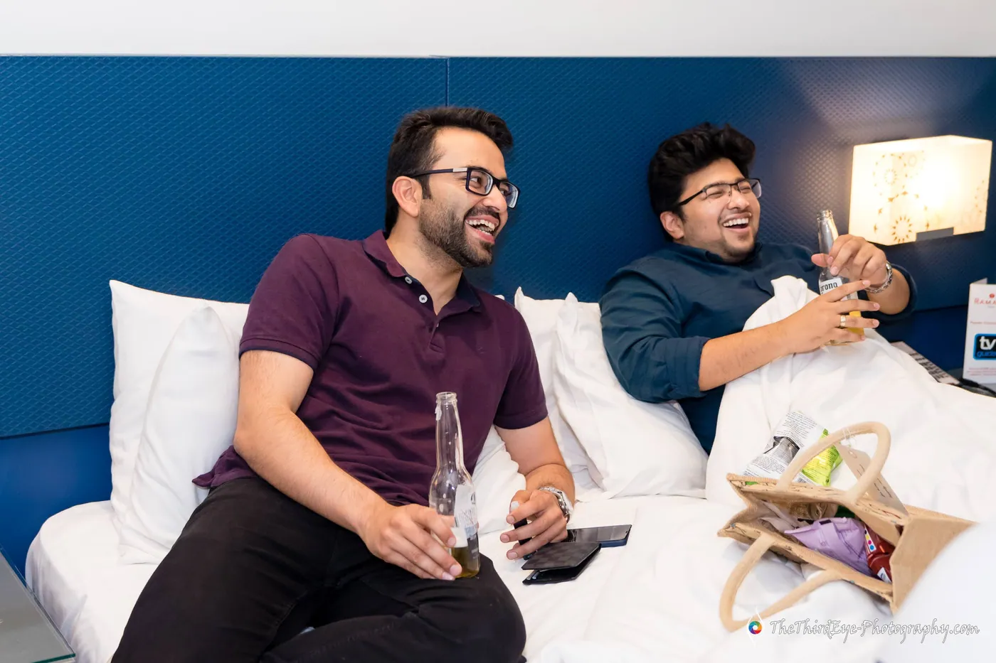 Two men laugh together on a hotel bed, one holding a beer bottle, a gift bag between them