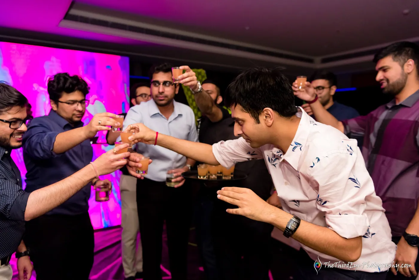 Friends lean in to clink shot glasses under a pink screen, the whole group laughing