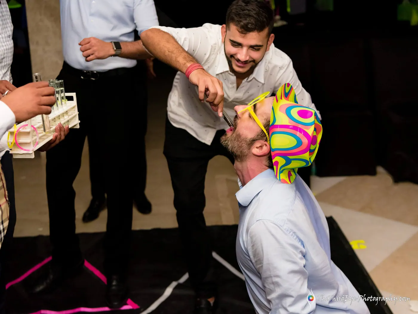 A man in a neon patterned hat tips his head back as a friend pours a shot