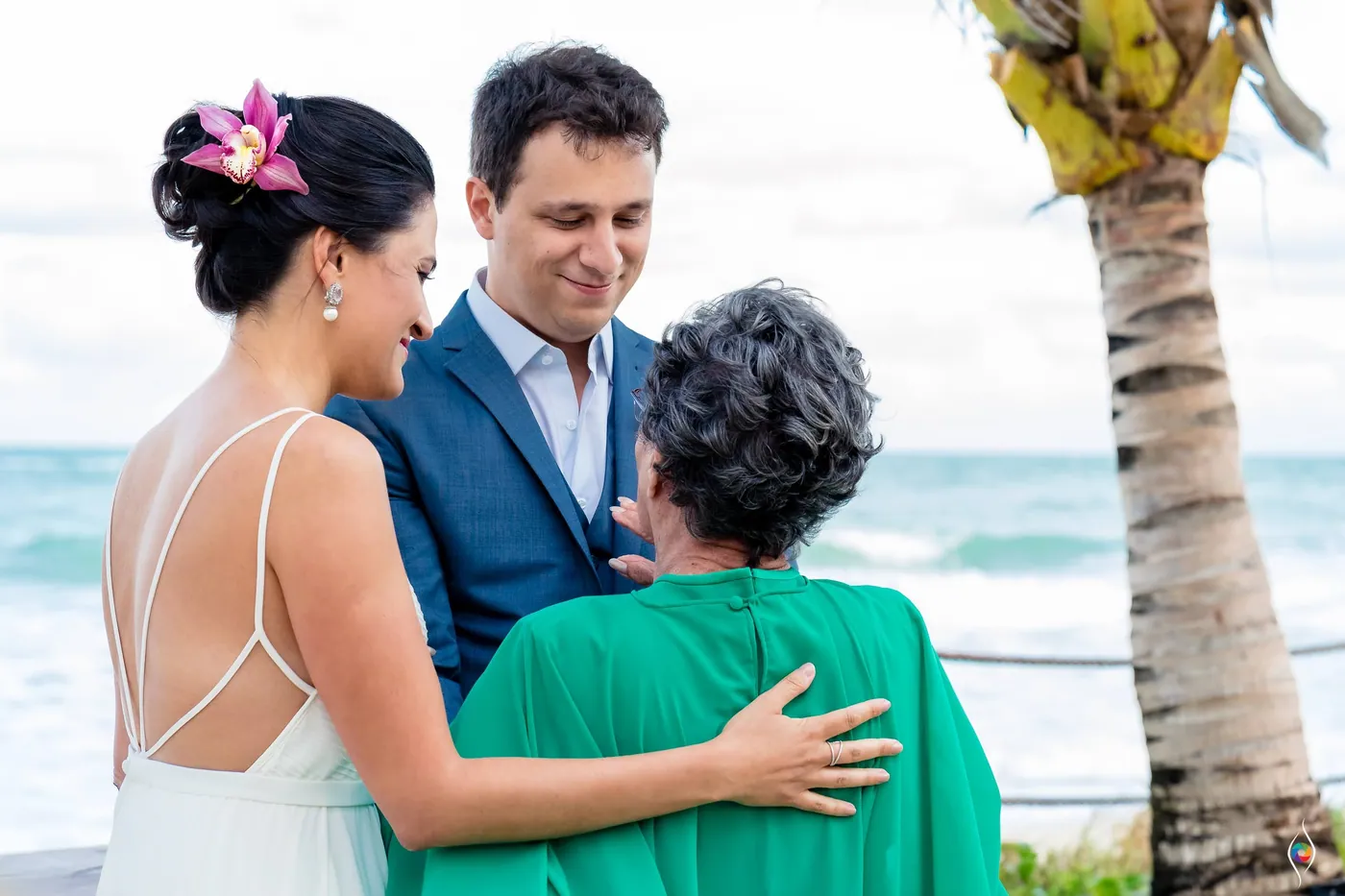 Evy hugs an older woman in green while Thiago smiles over the shoulder, palm and sea behind them