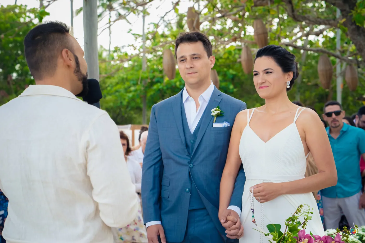 Evy and Thiago stand hand in hand facing a man in a cream shirt as guests look on