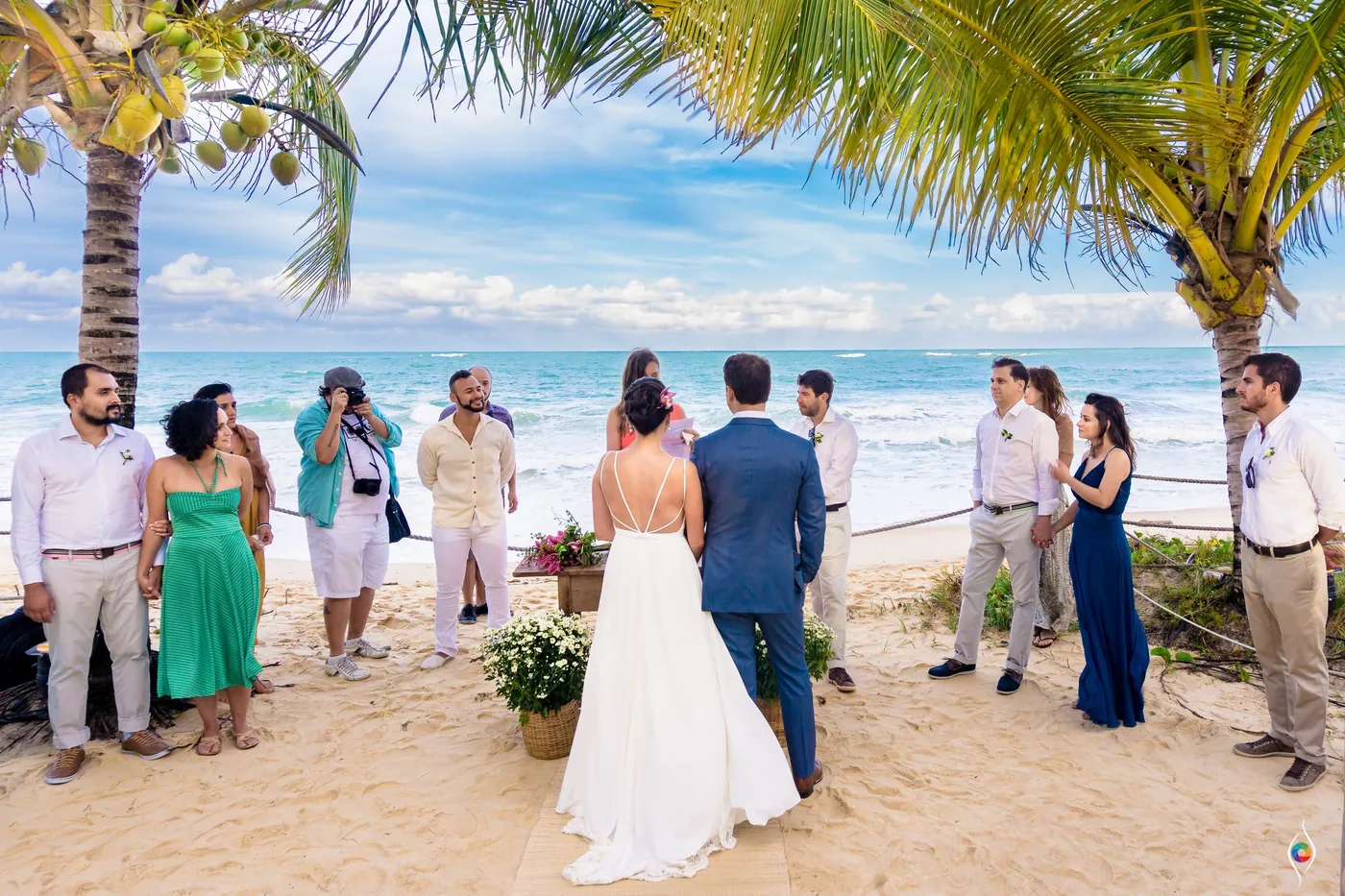 Seen from behind, Evy in her white gown stands with Thiago on the sand as guests watch beneath palms