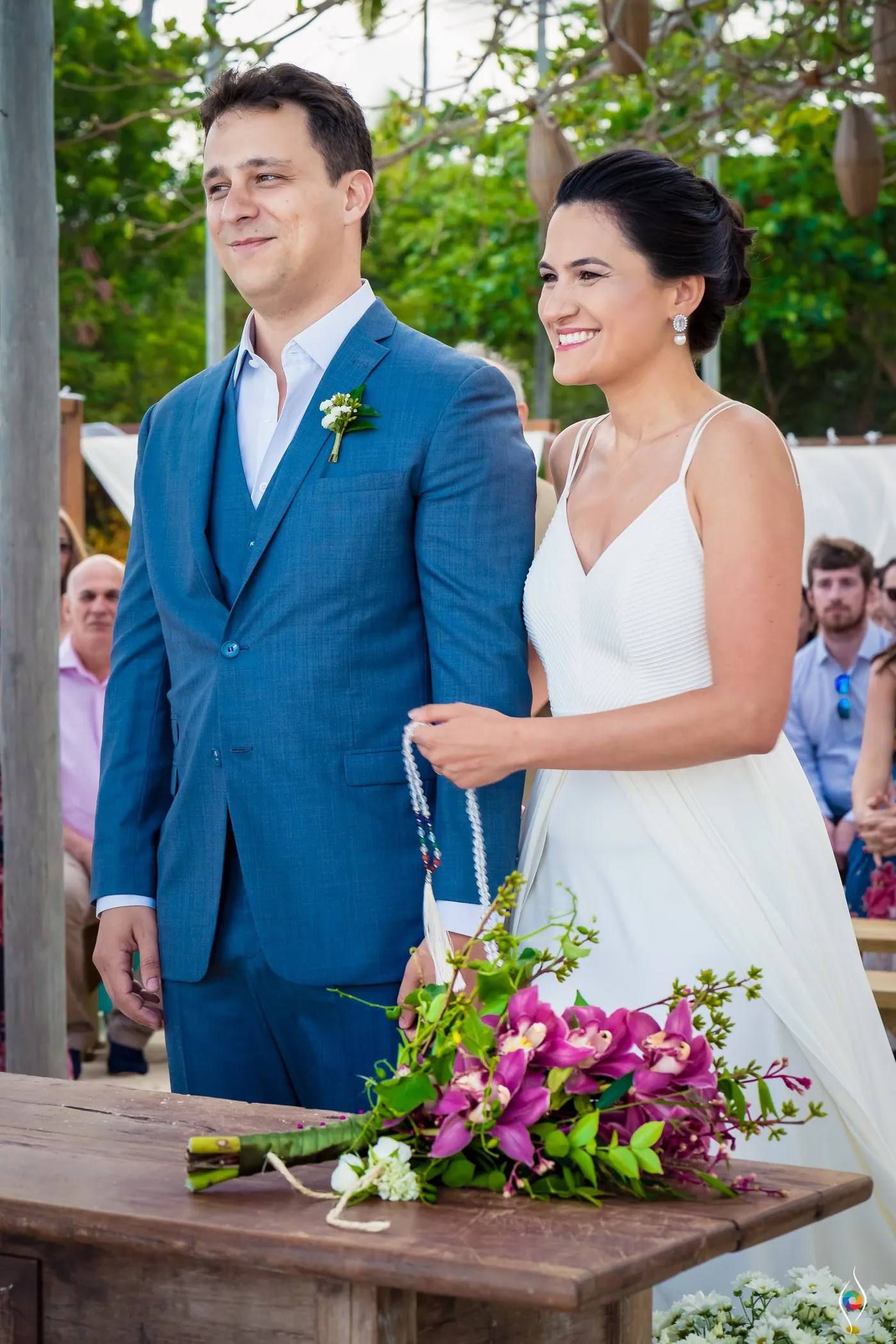 Thiago smiles off to one side while Evy looks down at their hands, pink flowers on the wooden table