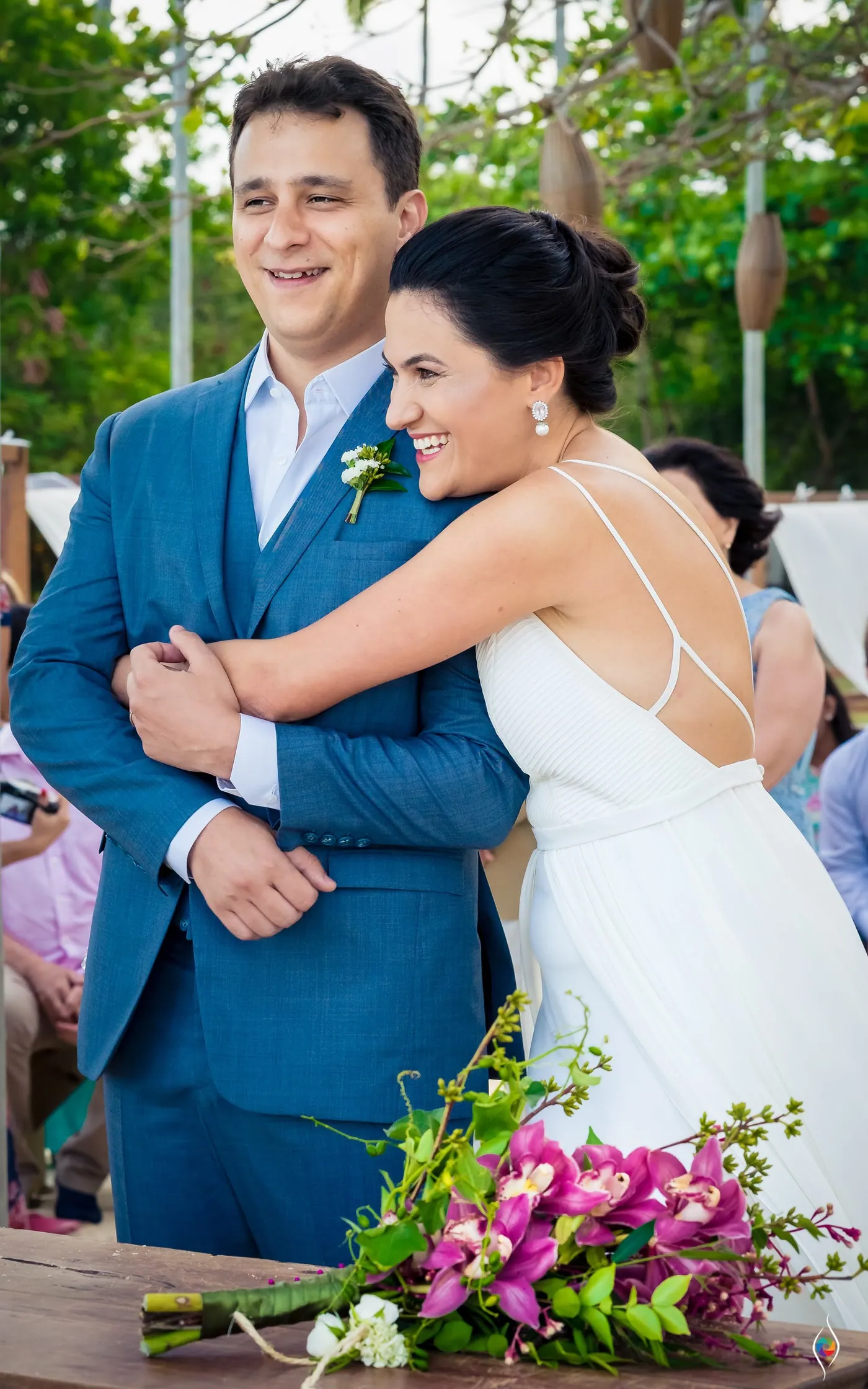 Evy wraps her arms around Thiago in a tight hug as he laughs, pink flowers on the table below