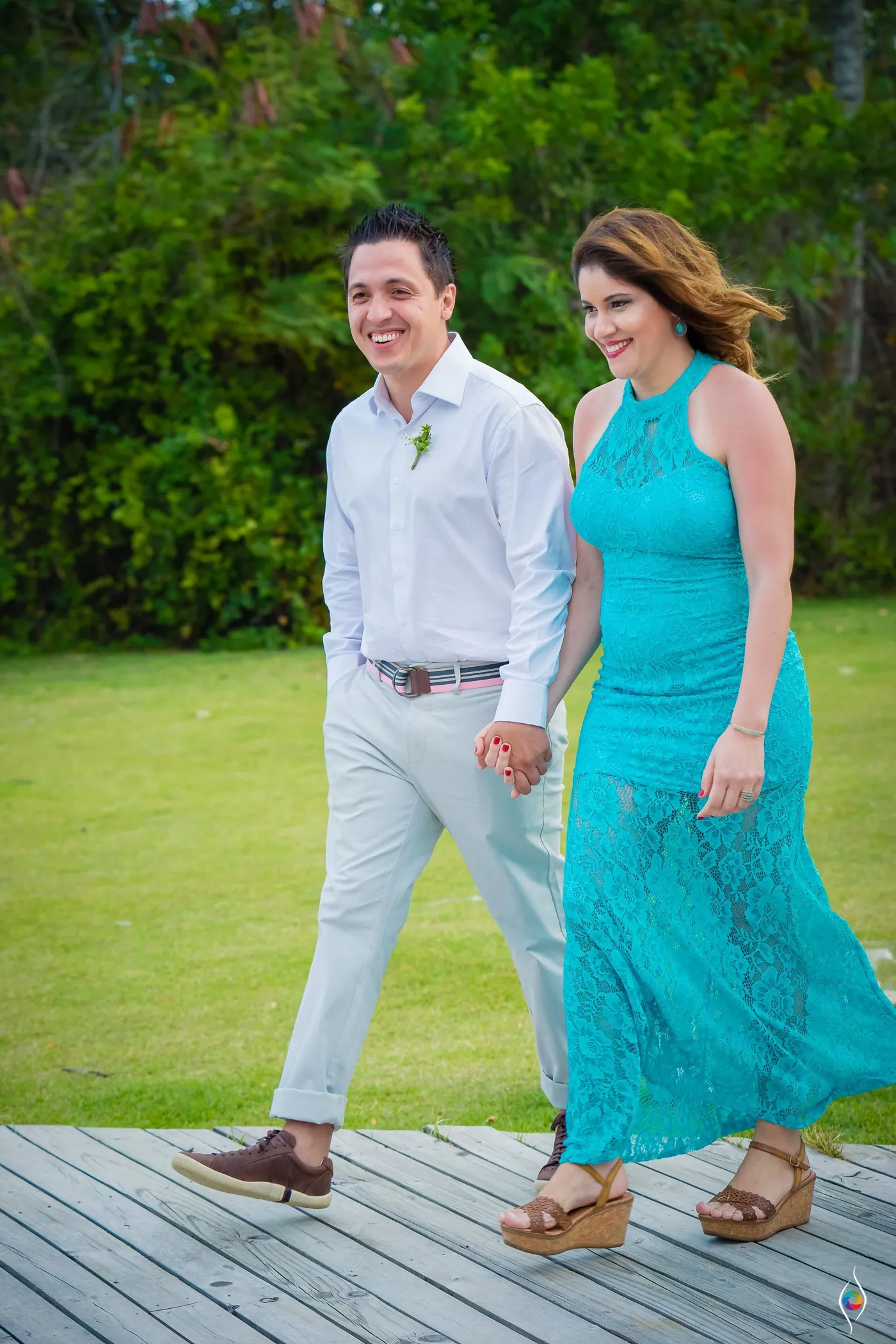 A couple in a teal lace dress walk hand in hand along a wooden deck