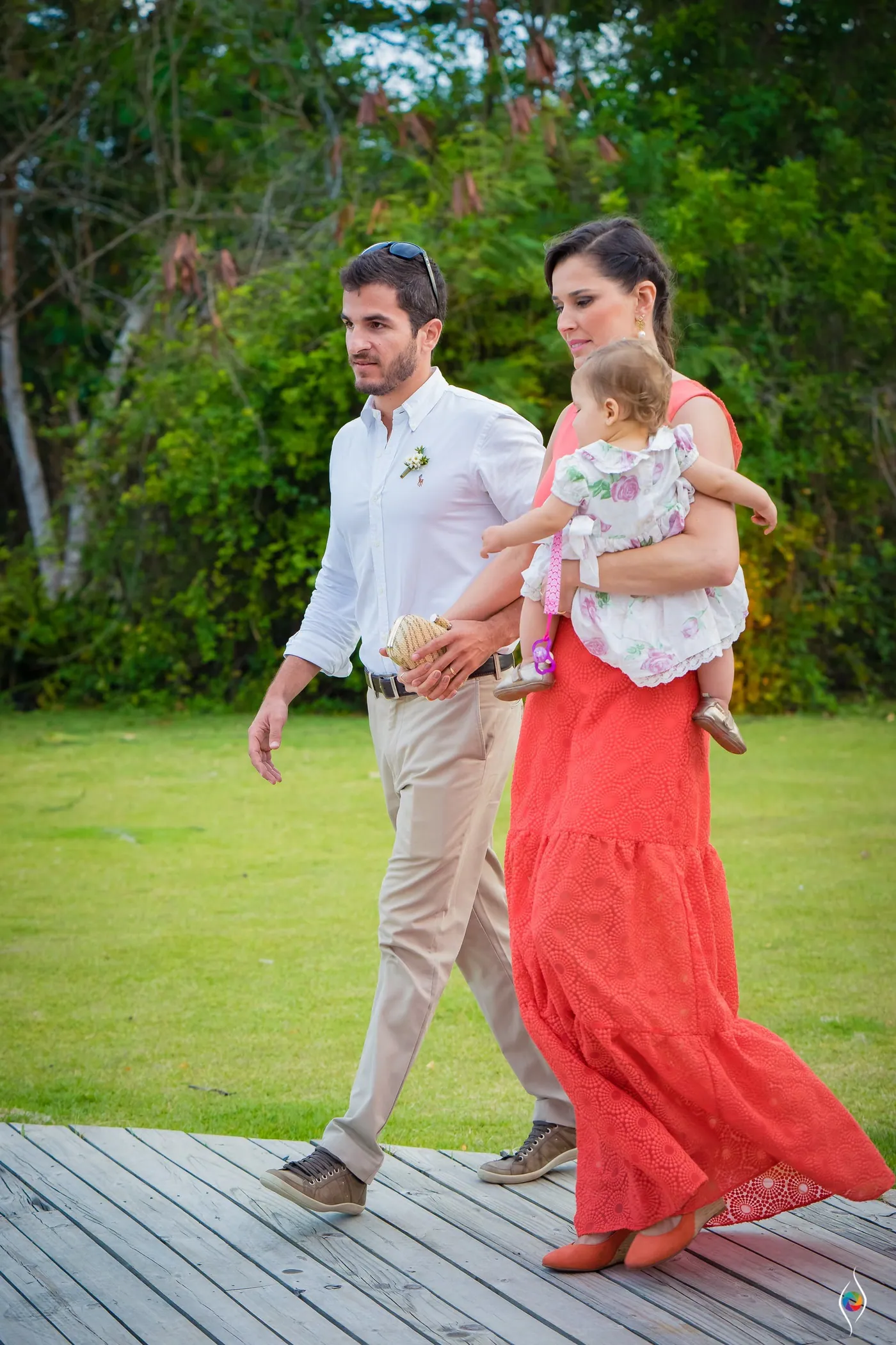 A man in white and a woman in a long coral dress carrying a baby cross a wooden deck